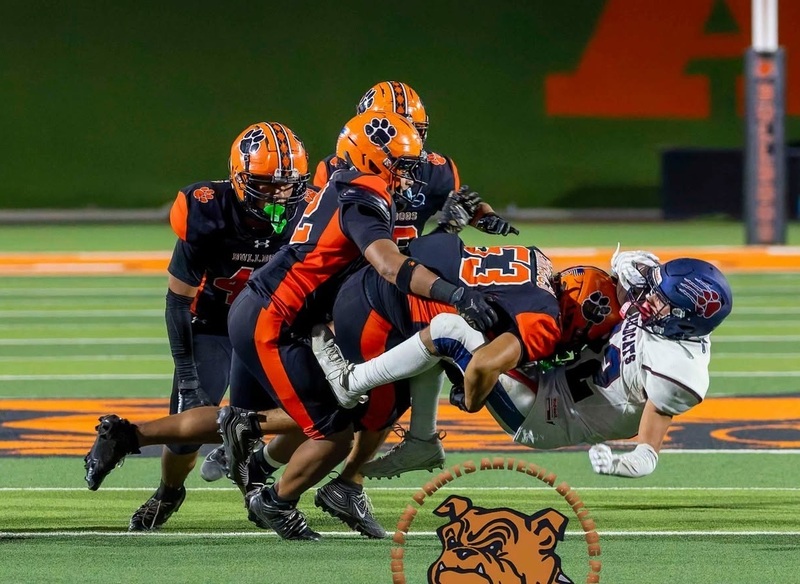 A teen boy in an orange and black football uniform knocks an opposing running back in white and blue off his feet with a powerful tackle as three teammates look on.
