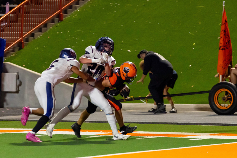 A teen boy in an orange and black football uniform leans forward as he carries the ball toward the goal line with a pair of defenders in white and blue attempting to tackle him.