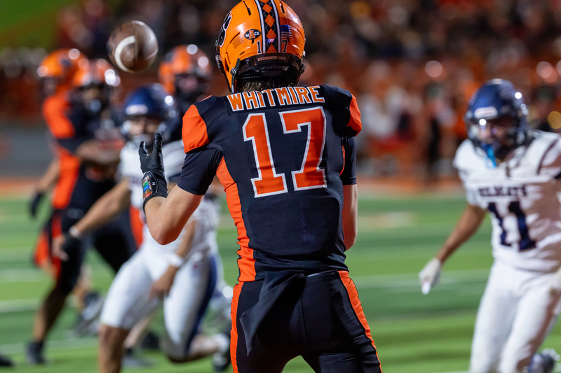 A teen boy in an orange and black football uniform with his back to the camera looks a mid-air football into his hands as opponents in white and blue look on.