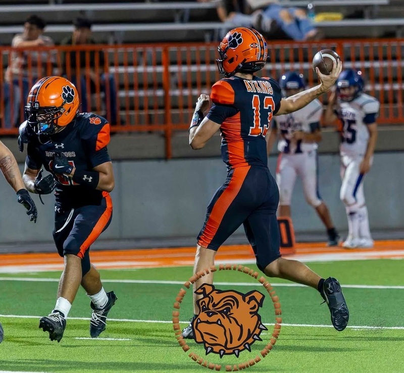 A teen boy in an orange and black football uniform rares his right arm back as he prepares to pass. A teammate in front of him looks to block.