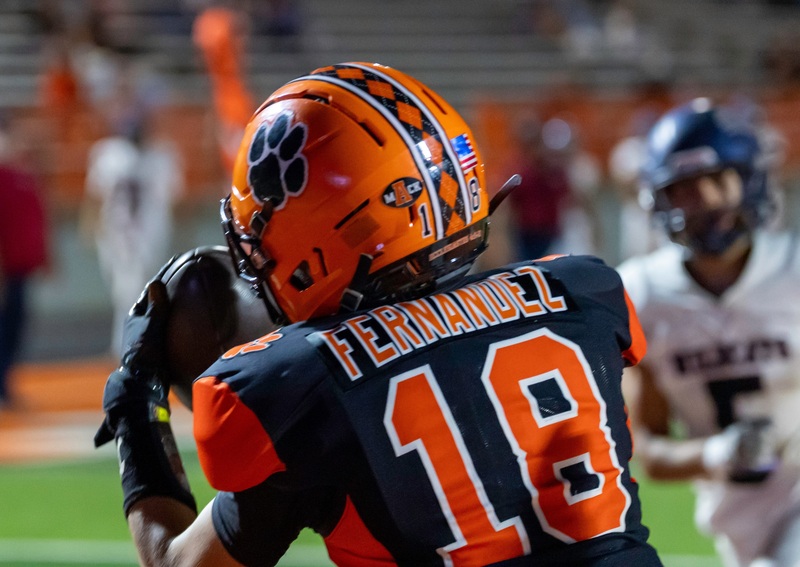 A teen boy in an orange and black football uniform with his back to the camera catches a football.