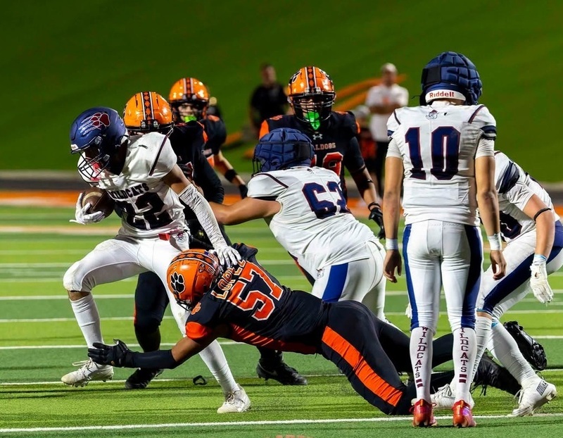 A teen boy in an orange and black football uniform dives forward to tackle the legs of a running back in white and blue.