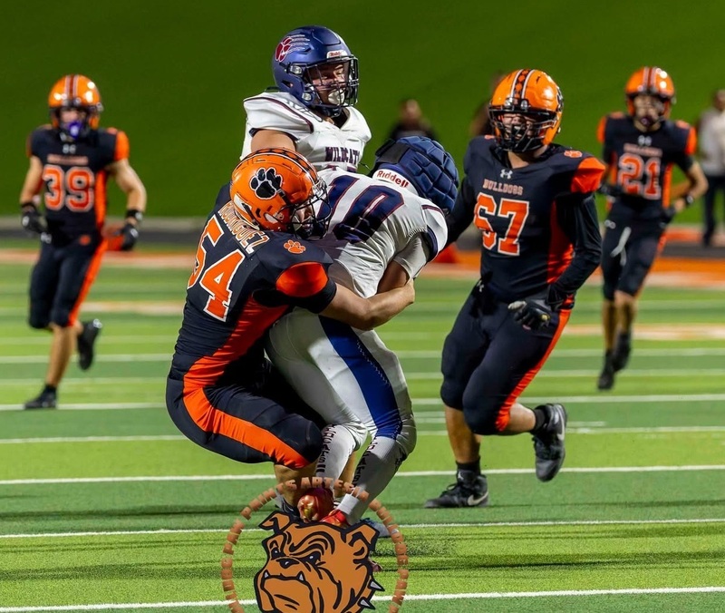 A teen boy in an orange and black football uniform sacks a quarterback in white and blue as teammates look on.