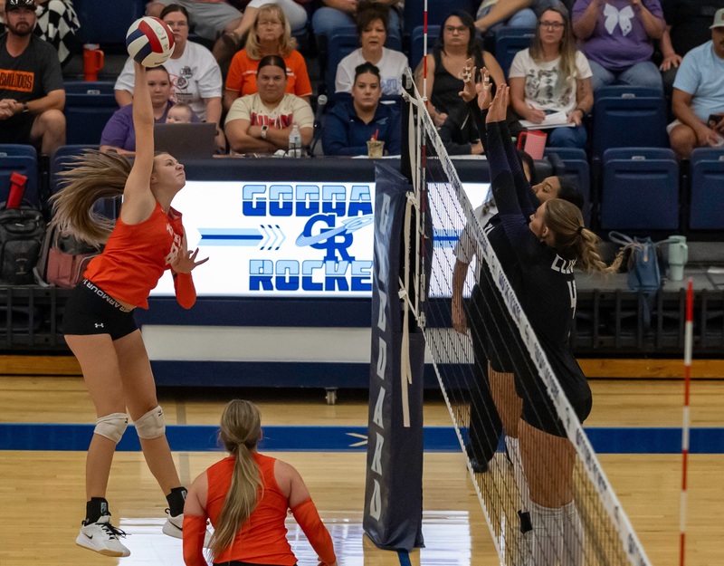 A teen girl in an orange and black volleyball uniform spikes the ball toward the net as opponents in black look to block.