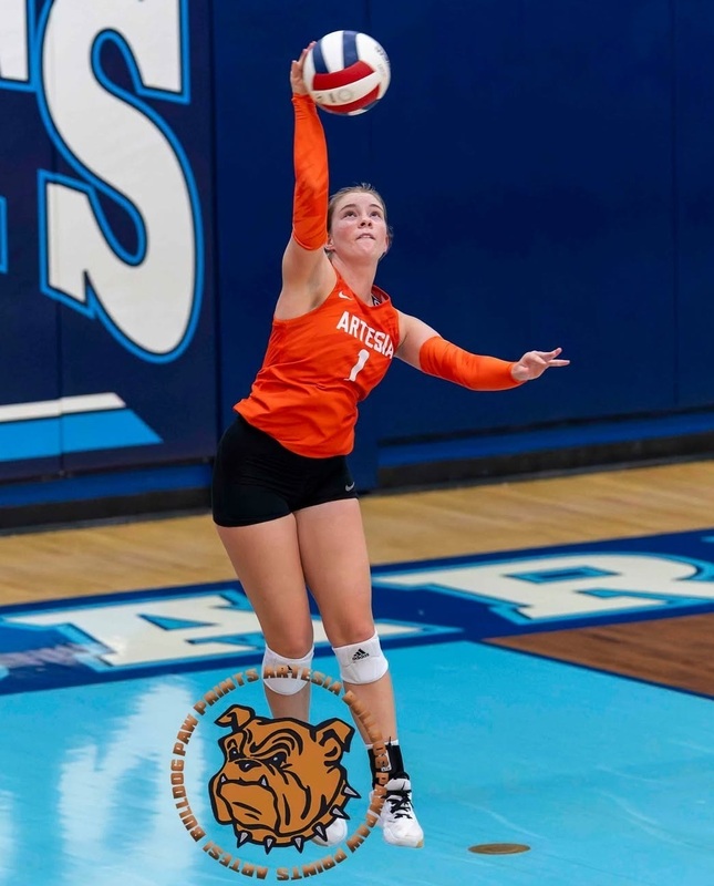 A teen girl in an orange and black volleyball uniform serves a red, white and blue ball as she jumps into the air.