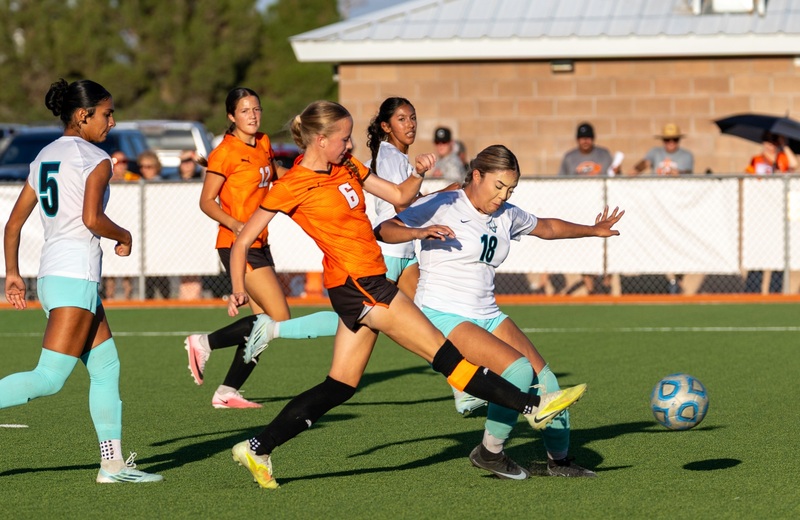 A teen girl in an orange and black soccer uniform kicks a ball with her right foot as an opponent in white and seafoam attempts to deflect the shot.