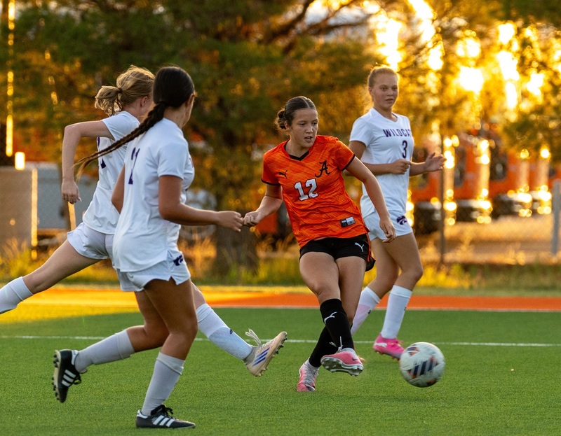 A teen girl in an orange and black soccer uniform kicks the ball with her right foot as three opponents in white surround her.