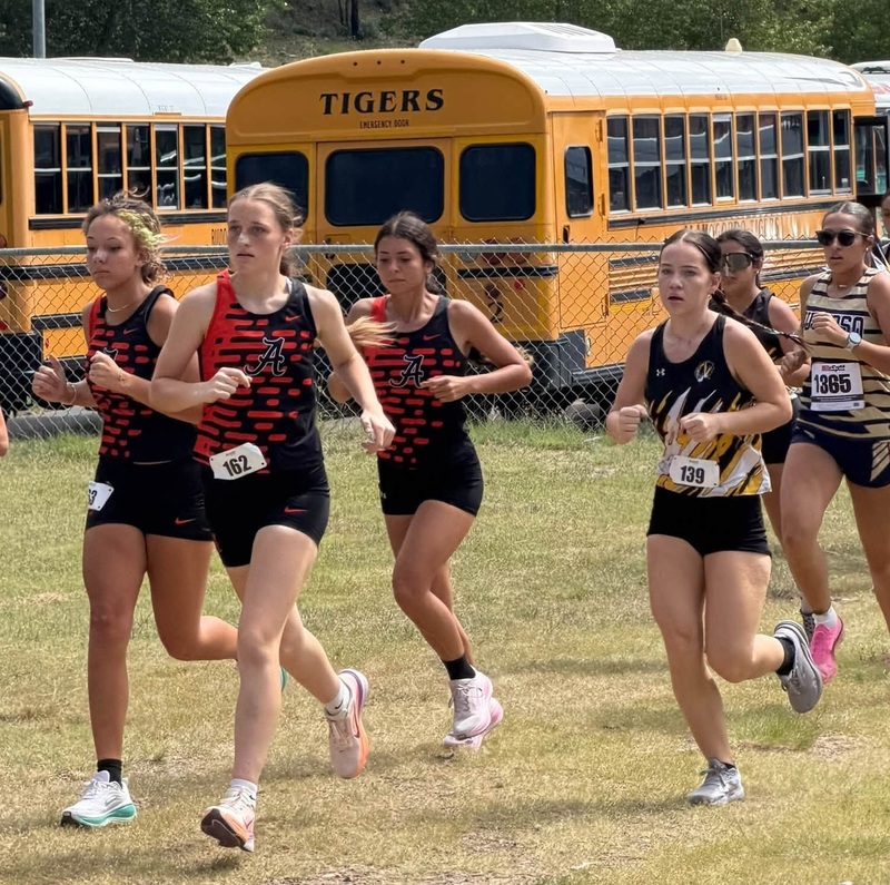 A group of teen girls in cross country uniforms run near the start of a race. Two buses are seen in the background.