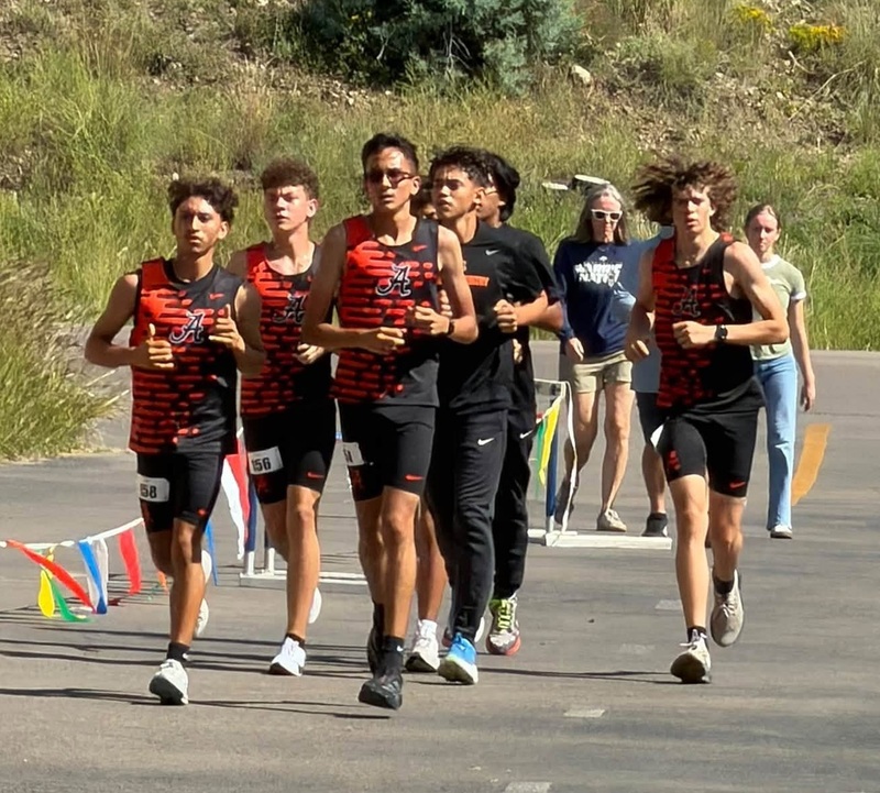 A group of teen boys in cross country uniforms run near the start of the race along a paved path.