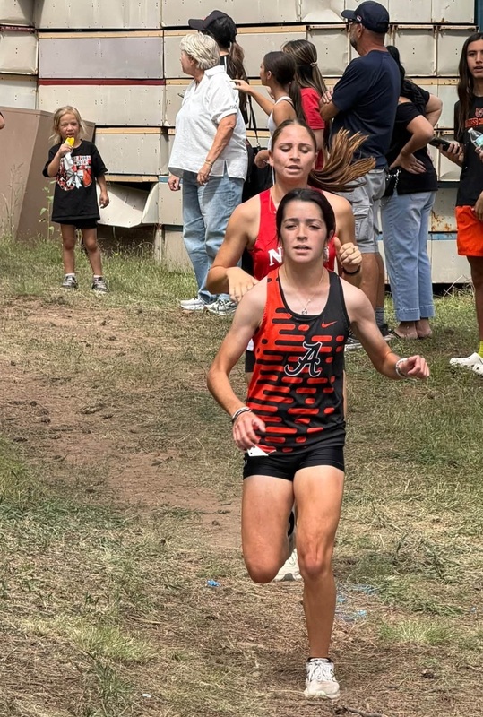 A teen girl in a black and orange cross country uniform runs down a grassy hill with an opponent behind her.