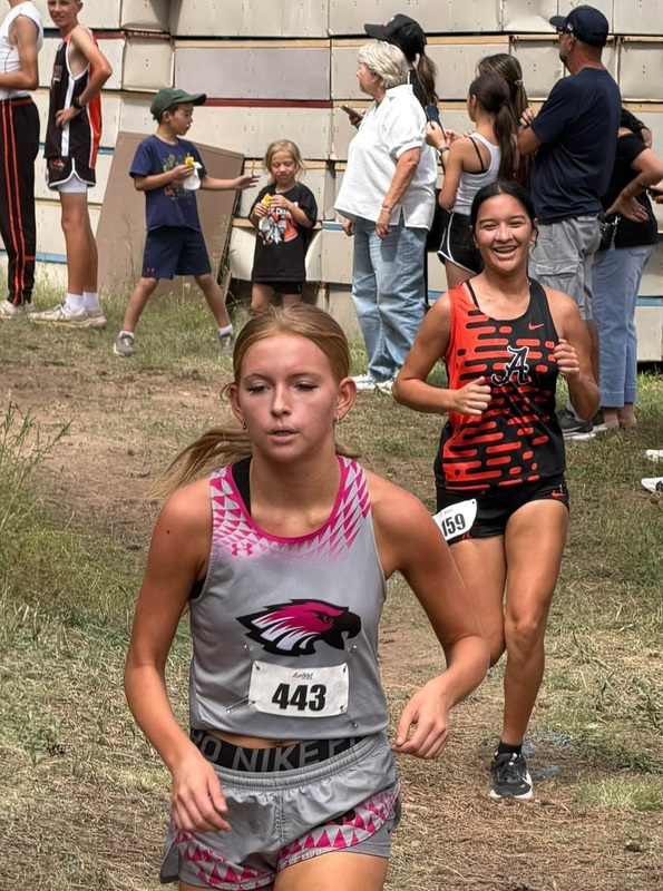 A teen girl in an black and orange cross country uniform smiles as she runs down a grassy hill behind an opponent in grey and red.
