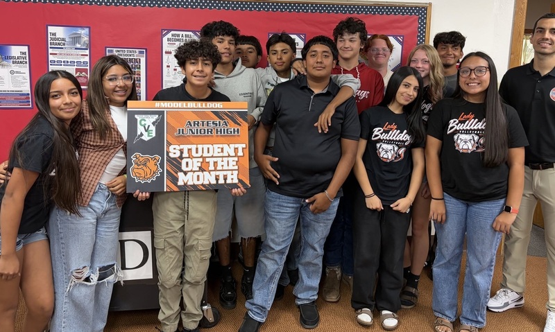 A teen boy holds a sign that reads "Student of the Month", surrounded by classmates and a male teacher.