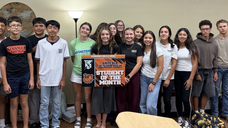 A teen girl holds a sign reading "Student of the Month", surrounded by classmates.