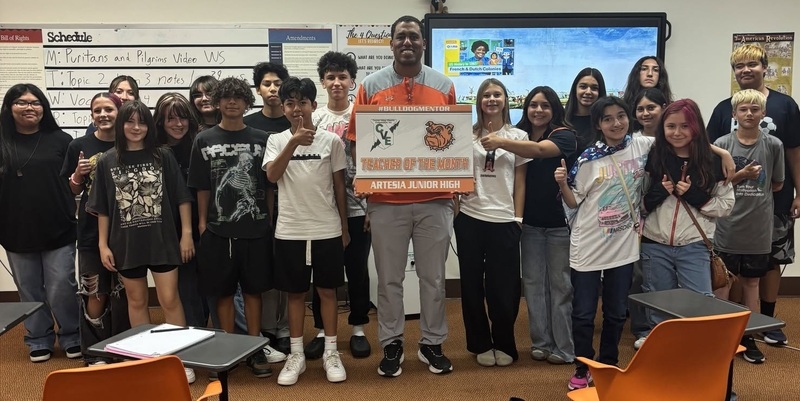 A male teacher holds a sign reading "Teacher of the Month", surrounded by students.