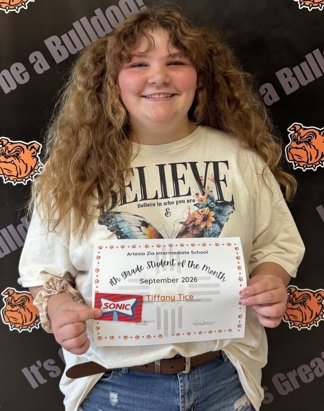 A preteen girl in a cream-colored t-shirt that reads "Believe" and jean shorts holds a certificate and a Sonic gift card as she smiles for the camera.