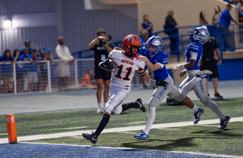 A teen boy in a white-and-orange football uniform steps over the goal line as he looks back at two players in blue-and-silver uniforms just behind him.