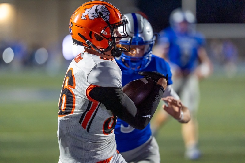 A teen boy in a white-and-orange football uniform catches the football just ahead of an opponent in blue and silver.