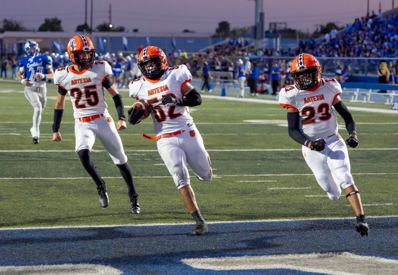 A teen boy in a white-and-orange football uniform runs into a blue end zone with the football. Two teammates run on either side of him.