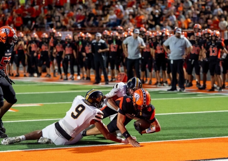 A teen boy in a black-and-orange football uniform dives over the goal line with the ball as two opponents in white and black hang from his torso. Behind him, the home sideline and stands can be seen.