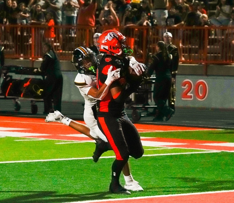 A teen boy in a black-and-orange football uniform runs across the goal line as an opponent in white and black hangs from his back.