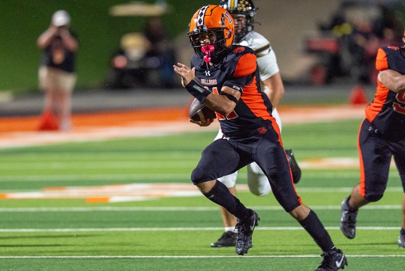 A teen boy in a black-and-orange football uniform runs with the football while looking ahead.