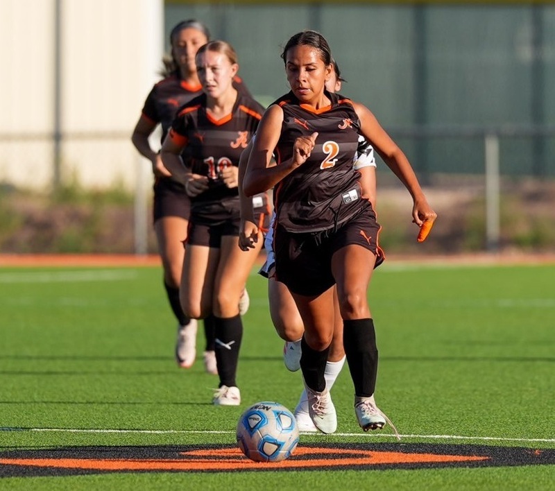 A teen girl in a black-and-orange soccer uniform dribbles the ball as two teammates follow behind her.