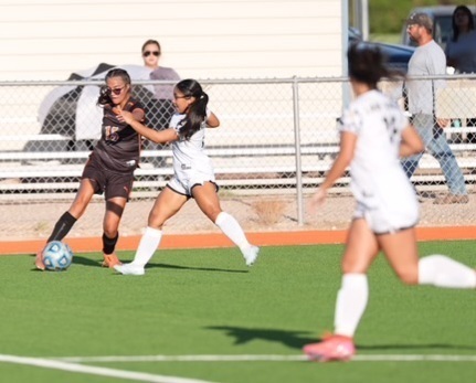 A teen girl in a black-and-orange soccer uniform kicks the ball away from an opponent in white.