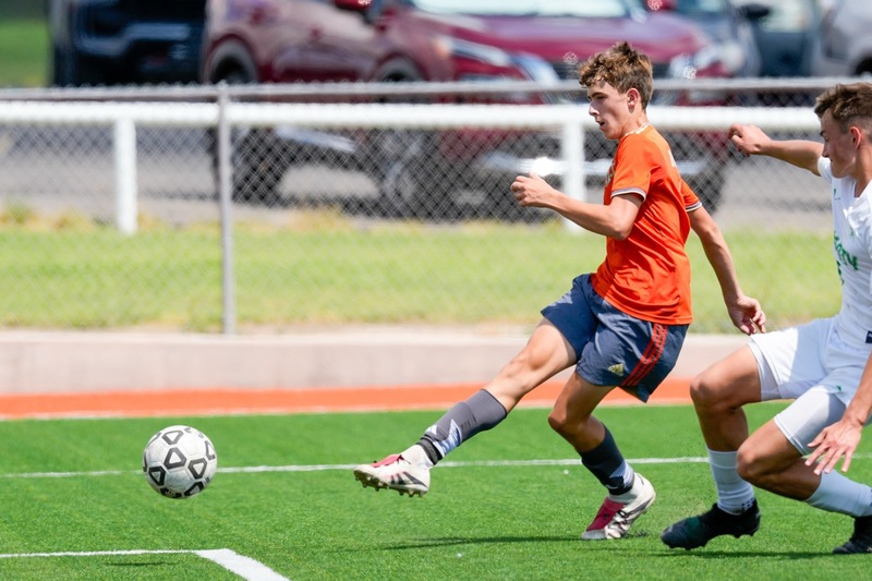 A teen boy in an orange-and-grey soccer uniform sends the ball into the air toward the goal as an opponent in white attempts to slide in.