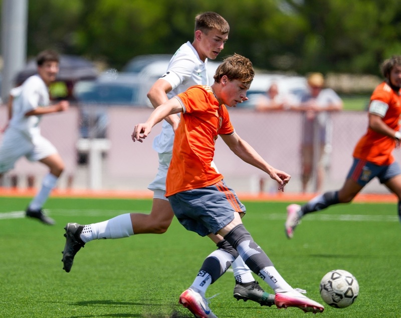 A teen boy in an orange-and-grey soccer uniform lunges to kick the ball as an opponent in white attempts to bump him off course.