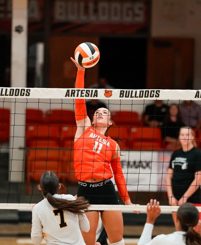 A teen girl in an orange-and-black volleyball uniform slaps an orange, black and white volleyball over a net.