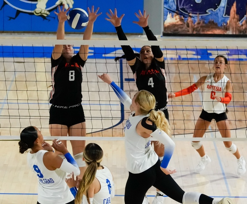 A teen girl in a black volleyball uniform blocks a blue and white volleyball back onto the opposing side of the net as three opponents in white and blue attempt to return it.