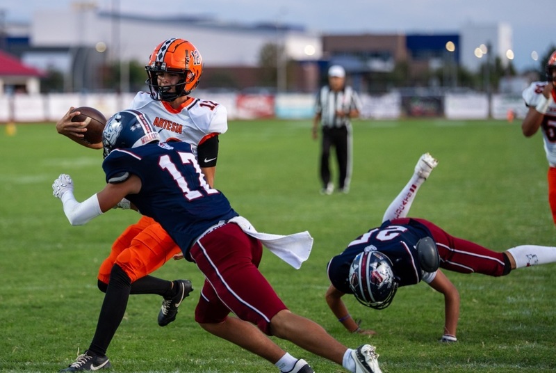 A teen boy in a white-and-orange football uniform runs with the ball in his right hand as two defenders in navy and maroon dive at him.