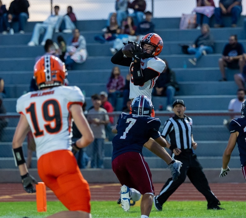A teen boy in a white-and-orange football uniform jumps into the air as he catches the ball with a defender in navy and maroon in front of him. 