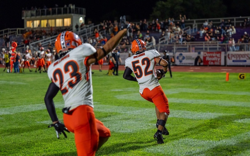 A teen boy in a white-and-orange football uniform runs through the end zone with the ball in his right arm as a teammate runs behind him with his arm raised in celebration.