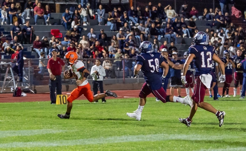 A teen boy in a white-and-orange football uniform sprints ahead of two defenders in navy and maroon.