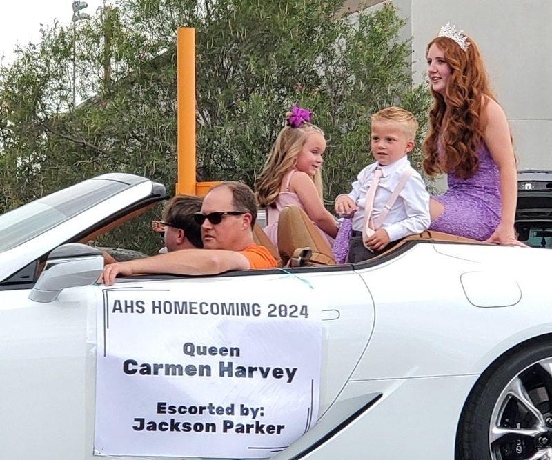 A teen girl in a gown and tiara sits on the back of a white car with two small children in front of her.