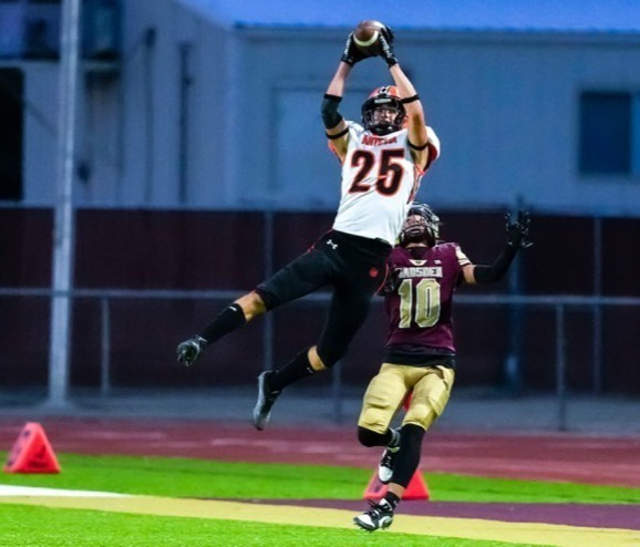 A teen boy in a white, orange and black uniform leaps high in the air to intercept a pass in front of an opponent in maroon and gold.