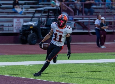 A teen boy in a white, orange and black uniform prepares to step into the end zone with the ball tucked into his right hand.