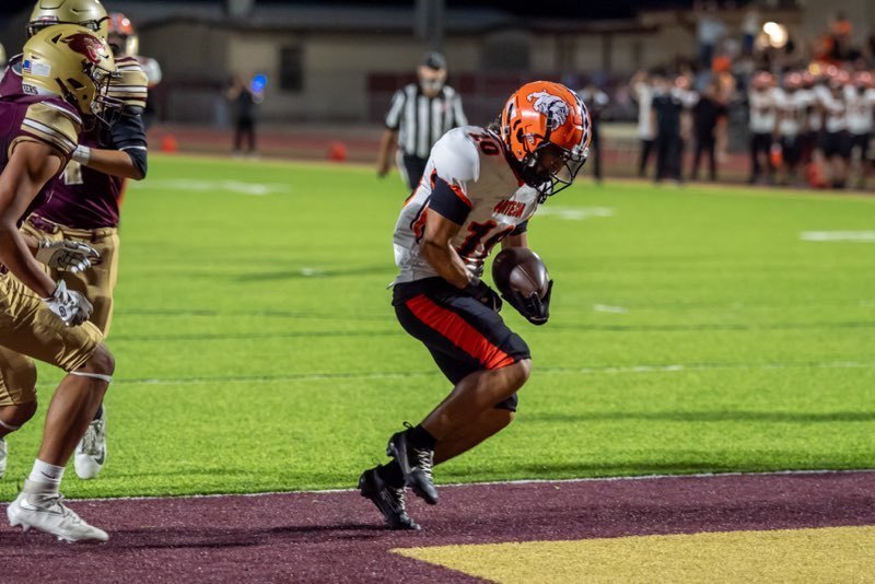 A teen boy in a white, orange and black uniform holds the ball curled into his left hand as he jumps into the end zone.
