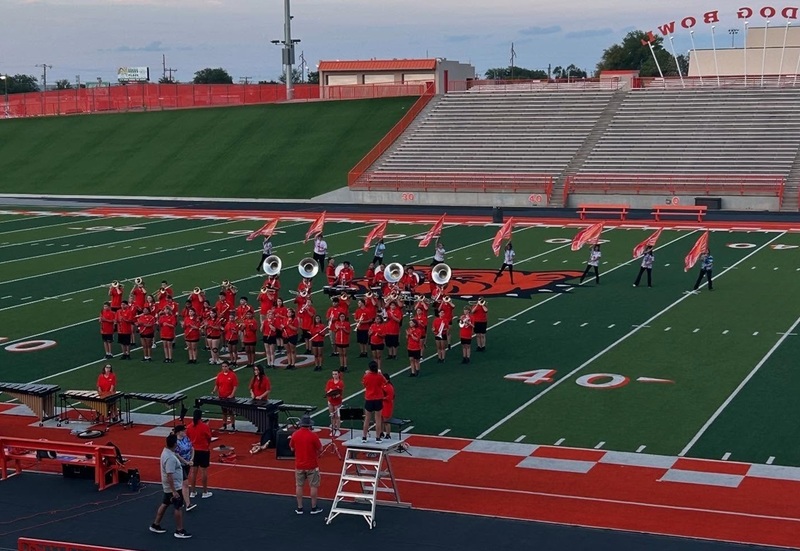 A high school marching band clad in orange t-shirts and black shorts marches across a football field.