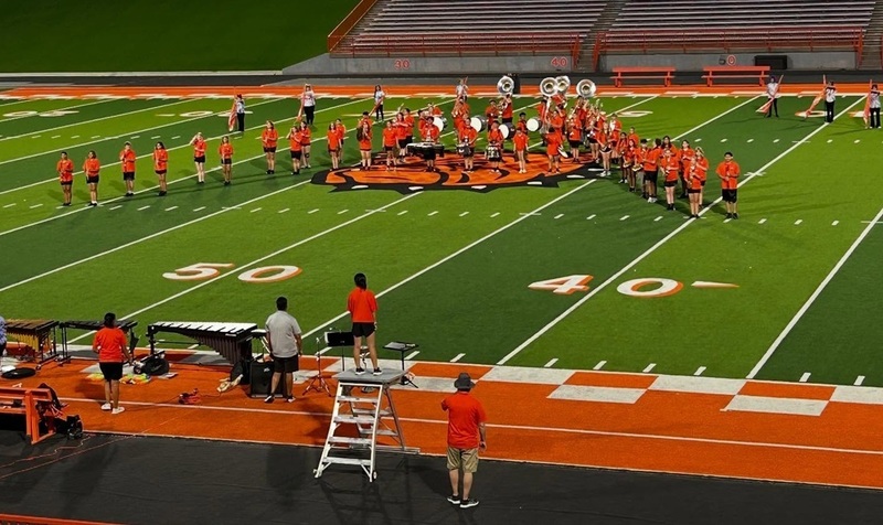 A high school marching band in orange t-shirts and black shorts plays the AHS fight song as they form an A on a football field.