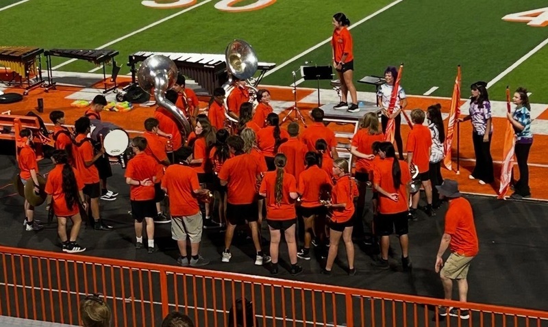 A high school marching band in orange t-shirts and black shorts stands on the track adjacent a football field holding their instruments.