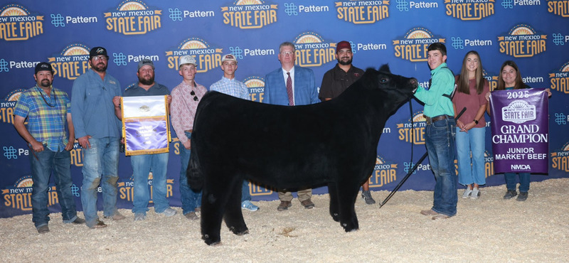A teen boy in a turquoise shirt and jeans holds the head of a large black steer as men and women stand behind him, some holding banners.