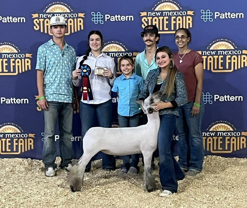 A teen girl in jeans and a sparkly silver shirt holds the head of a lamb as family members stand behind her holding a ribbon and a buckle.