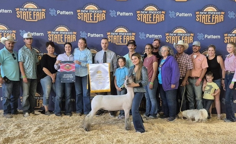 A teen girl in jeans and a sparkly silver shirt holds the head of a lamb as multiple people stand behind her, two holding banners.