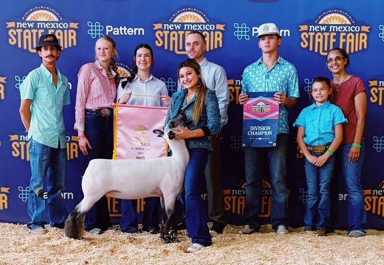 A teen girl in jeans and a sparkly silver shirt holds the head of a lamb as family members stand behind her, two holding banners.