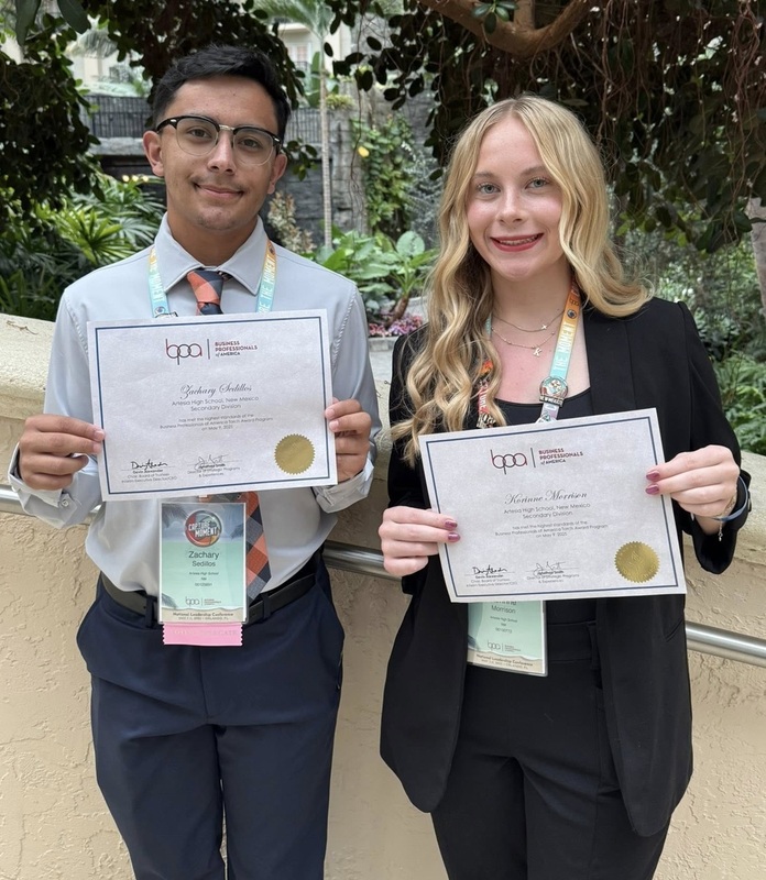 A teen boy and teen girl smile for the camera as they hold certificates.