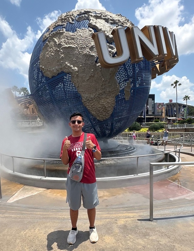 A teen boy in a red shirt and shorts gives a thumbs-up as he stands in front of a Universal Studios globe.