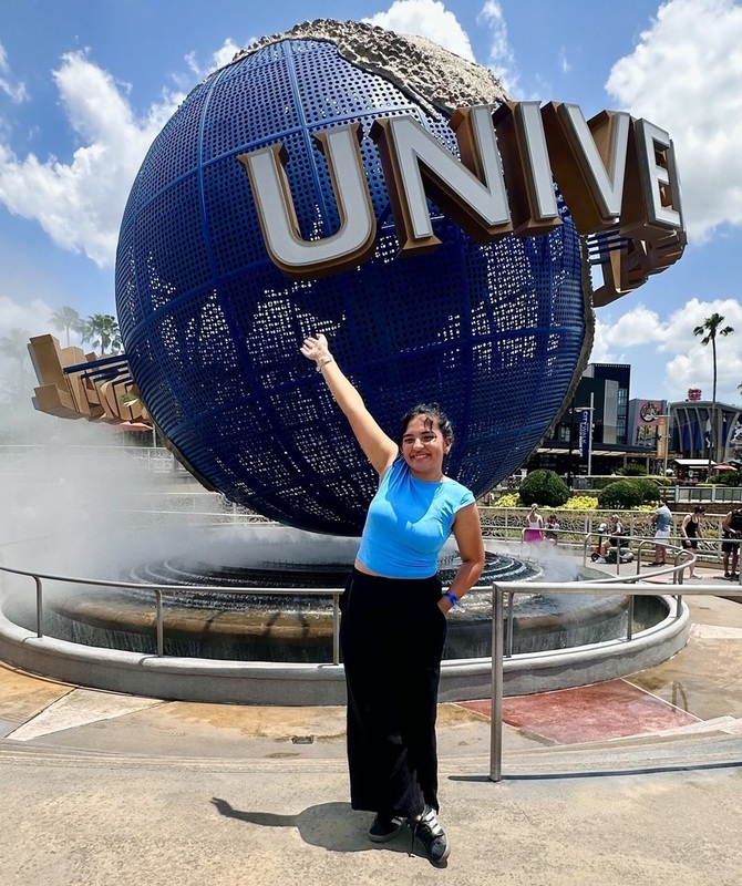 A teen girl in a blue shirt and black pants raises her right arm in the air as she stands in front of a Universal Studios globe.
