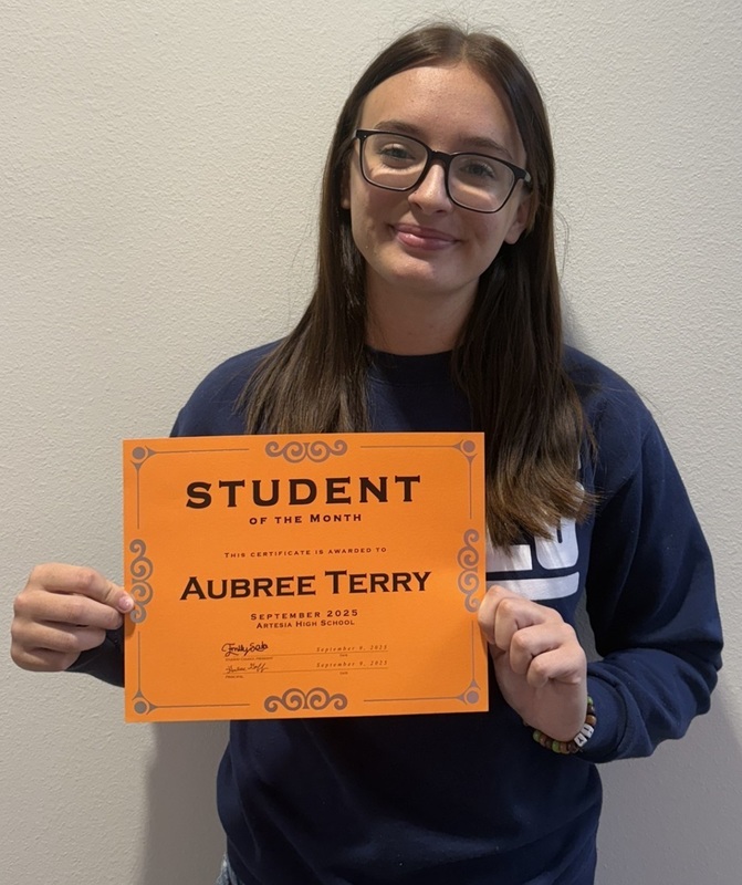 A teen girl in a navy long-sleeved shirt and glasses holds an orange sign reading: "Student of the Month: Aubree Terry".